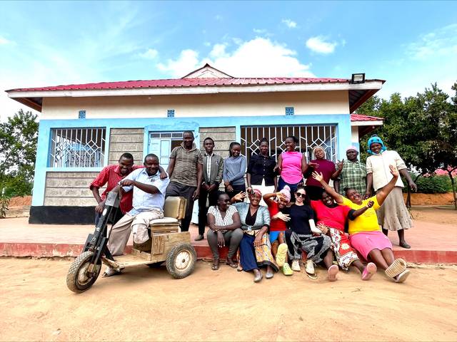 A group of happy people posing in front of a house with one person on a motorcycle.