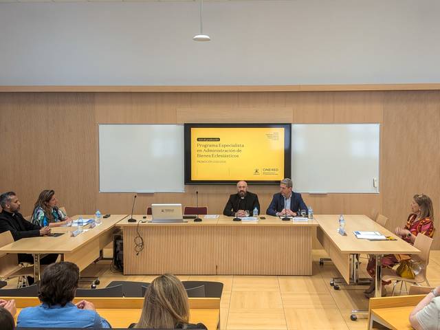 Una reunión académica en un aula con varias personas sentadas alrededor de una mesa, escuchando a los ponentes.
