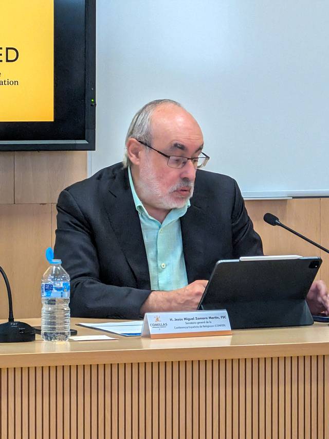 Un hombre de cabello canoso y barba, vestido con un traje oscuro, está sentado frente a una mesa con un dispositivo electrónico, concentrado en su trabajo.