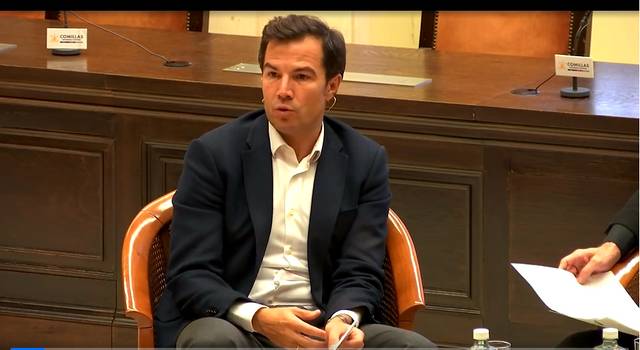 Un hombre sentado en una silla dentro de una sala de conferencias, vestido con traje oscuro y camisa blanca, parece estar escuchando atentamente.