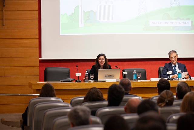 A woman is speaking at a conference while a man sits beside her, in front of an audience.