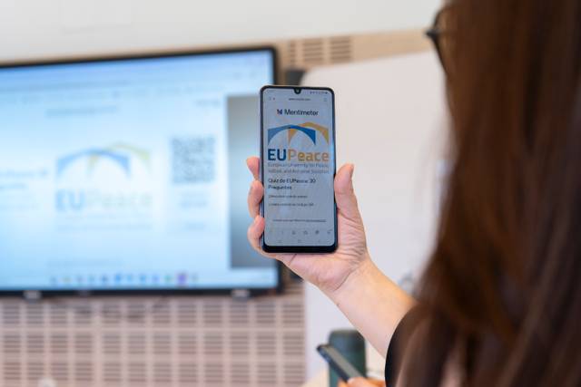 Una persona sostiene un teléfono móvil mostrando el logo de EUPeace mientras hay una pantalla grande detrás.
