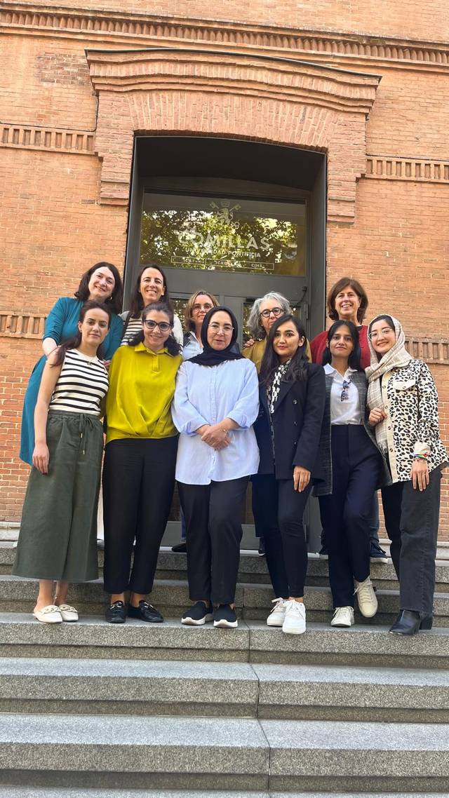 A group of nine women of diverse appearances standing on the steps of a brick building.