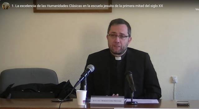 A priest giving a lecture in a classroom setting with a presentation titled '1. The excellence of Classical Humanities in the Jesuit school in the first half of the 20th century'.