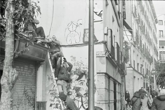 Un grupo de personas trabaja en la parte exterior de un edificio, con una escalera y grafitis visibles en la pared.