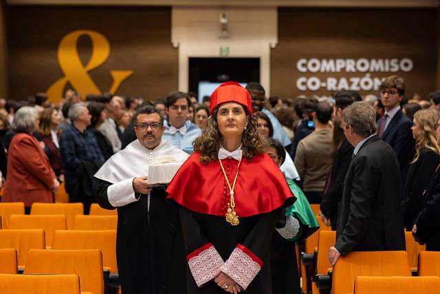 Ceremonia académica con un grupo de personas en un auditorio, destacando a una mujer con símbolo de autoridad en primer plano.