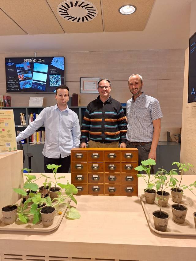Tres personas posando frente a un mueble con plantas y un panel informativo detrás.