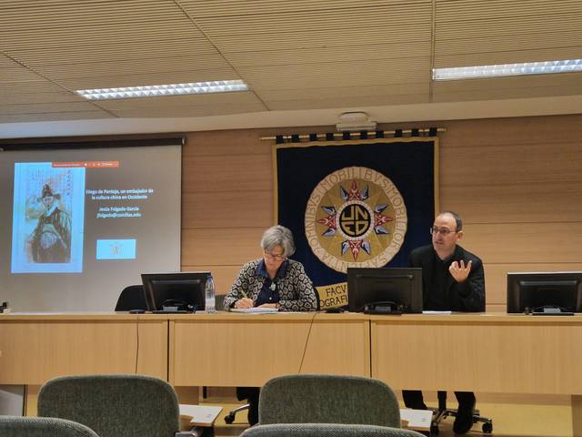 Dos personas sentadas en una mesa durante una presentación, con un proyector mostrando una imagen detrás de ellas.