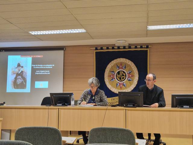 Dos personas están haciendo una presentación en una sala de conferencias con un escudo y una insignia de la UCM en el fondo.