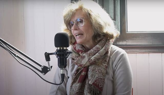 A woman with blonde hair wearing glasses and a scarf speaks into a microphone in a room with wooden paneling.