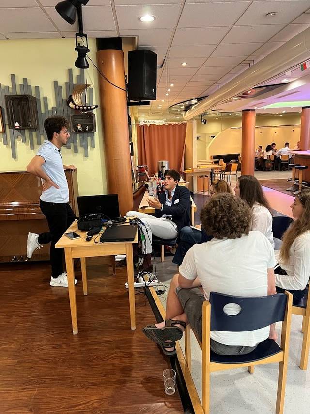 A group of people sitting and listening to a man who is standing and speaking in a casual indoor setting.