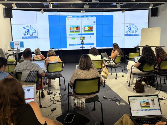 Un grupo de personas asiste a una presentación en una sala equipada con pantallas y computadoras portátiles.