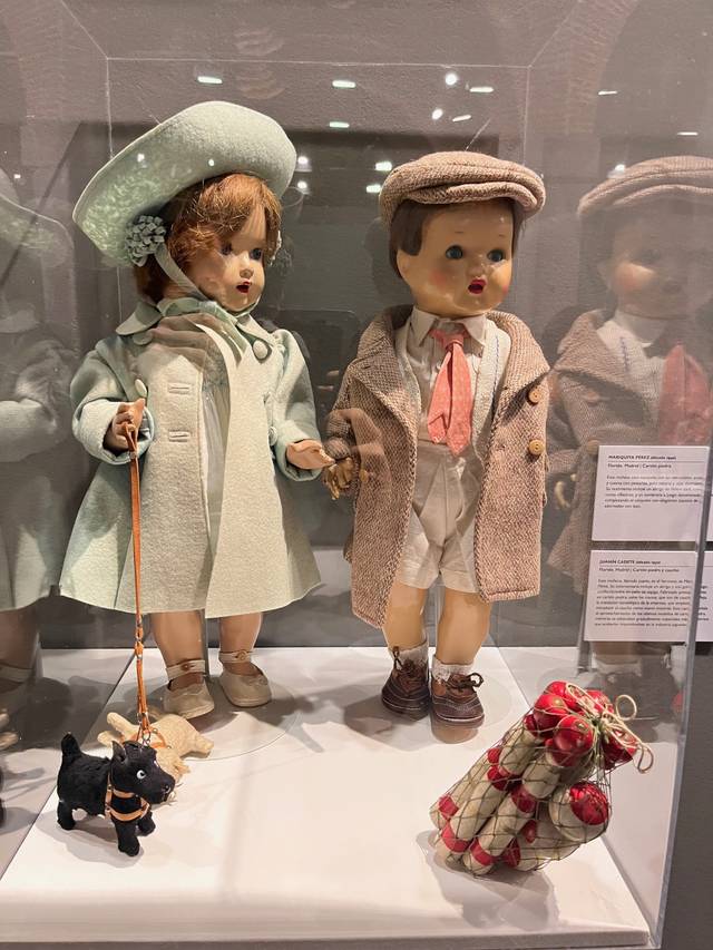 Dos muñecos vestidos con ropa de estilo antiguo están de pie en una vitrina, uno de ellos sostiene una correa con un perro de juguete y el otro está junto a un saco de otros muñecos pequeños.