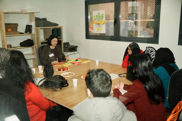 Un grupo de personas se reúne en una sala de trabajo, con una mujer liderando la conversación y una mesa llena de frutas y notas adhesivas.