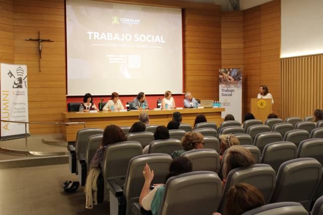 A person standing at a podium presenting to an audience in a conference room, with a panel seated behind them and a slide about Social Work displayed.