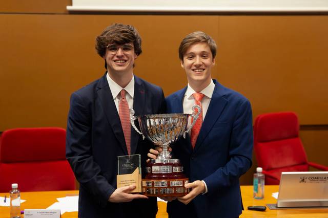 Dos jóvenes sonrientes vestidos con trajes formales sostienen un gran trofeo en una sala de conferencias.