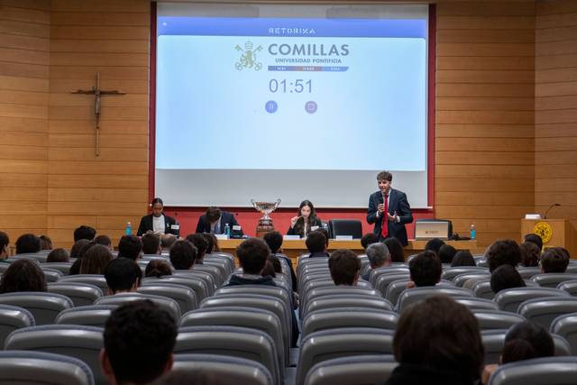 Un evento académico en la Universidad Pontificia Comillas con un ponente hablando frente a un público sentado.