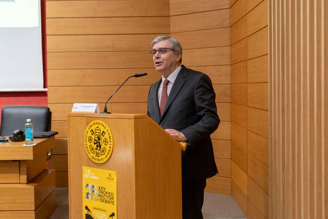 Un hombre habla desde un podio en un evento formal con paneles de madera en el fondo.