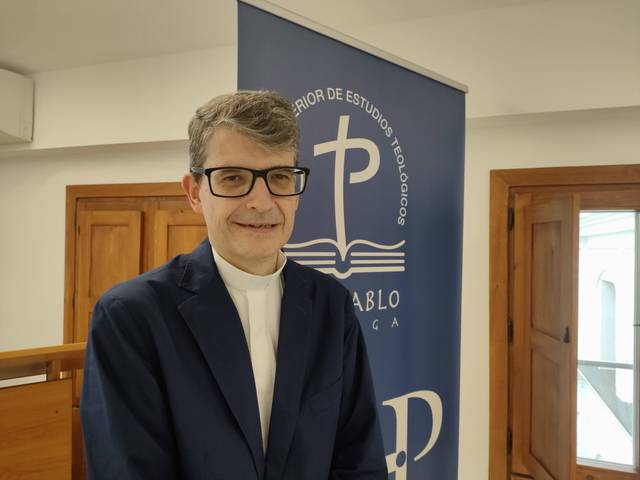 A man in a blue suit and glasses standing in front of a banner with educational or institutional logos.