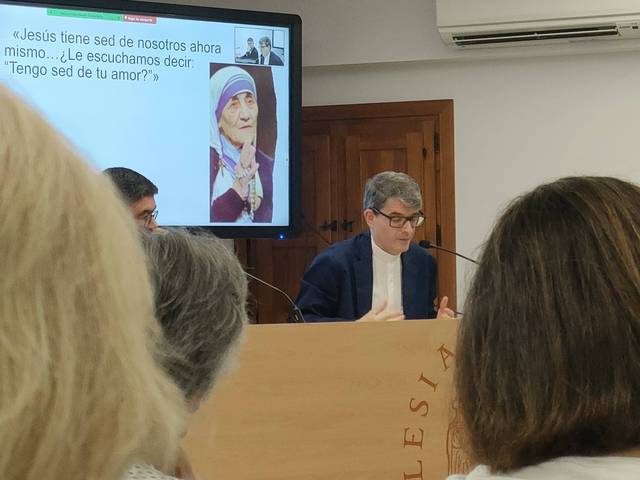 A man is speaking at a podium in a church meeting, with a presentation behind him featuring an image of Mother Teresa.