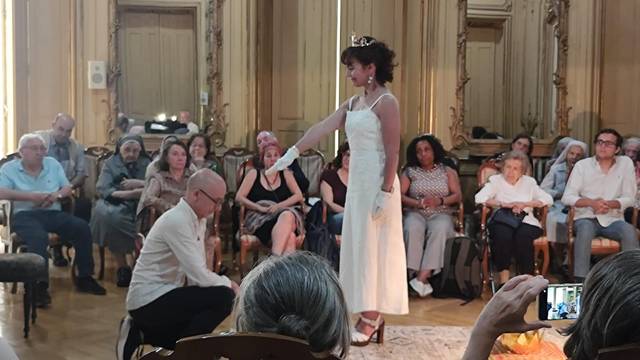 A woman in a white dress stands while a man kneels in front of her in an ornate room filled with an audience.