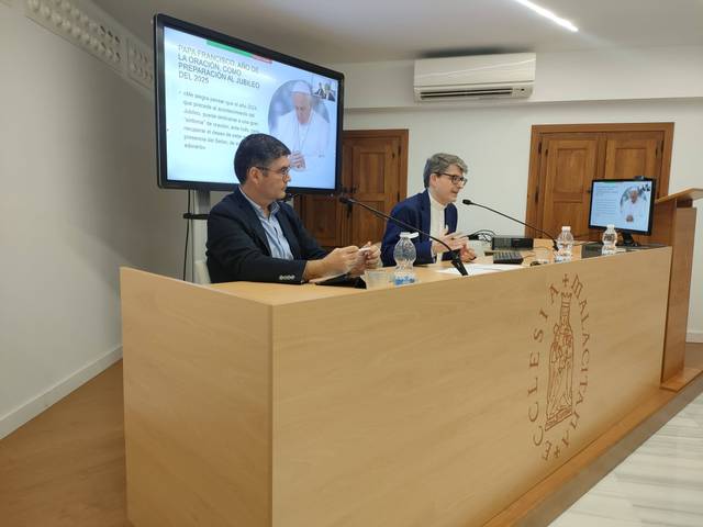 Two men sitting at a podium in a seminar room, presenting with a slideshow in the background.