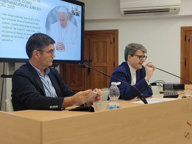 Two people are seated at a table discussing, with a presentation slide of Pope Francis in the background.