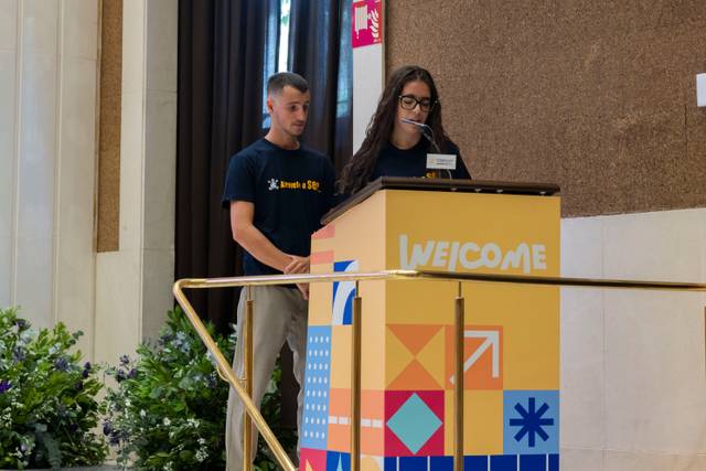 Dos personas están en un podio dando la bienvenida en un evento.