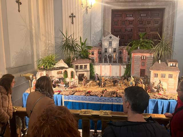 Un belén navideño decorado con figuras y edificios, mientras varias personas lo observan con atención.