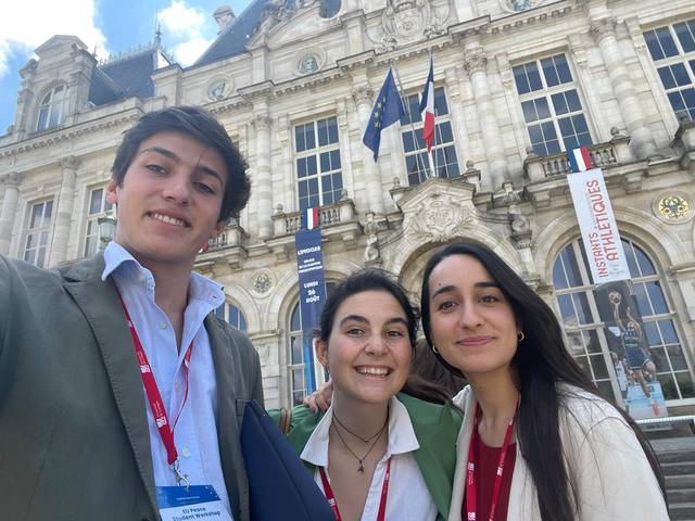 Three young adults are smiling for a selfie in front of a historic building with flags.