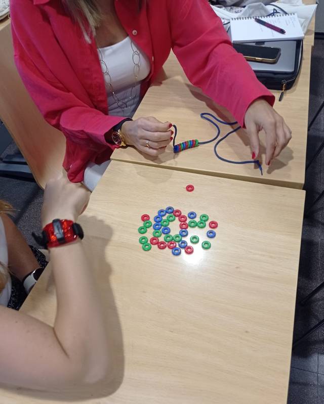 A person works on an activity involving multicolored round tokens on a wooden table, with another individual partially shown assisting.