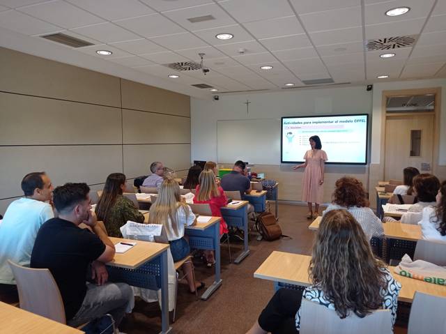 A woman is presenting to a group of adults seated at desks in a classroom setting.