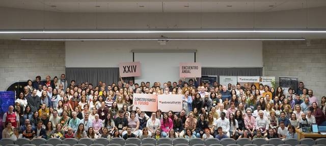 A large group of people of various ages holding a banner at an indoor event, with many smiling at the camera.