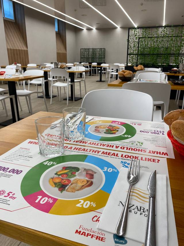 A view of a cafeteria table with promotional nutritional placemats and dining utensils, highlighting a healthy meal guide, set against a backdrop of empty white tables and chairs.