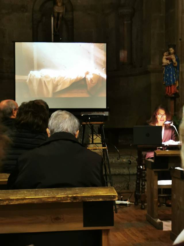 A woman presents a lecture indoors with a projected image behind her, in a room that appears to be a church.