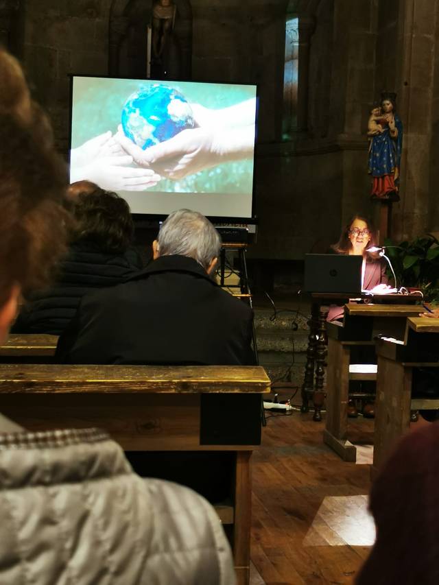 A woman sits at a desk in a church giving a presentation, a projected image of hands holding a small globe visible in the background.