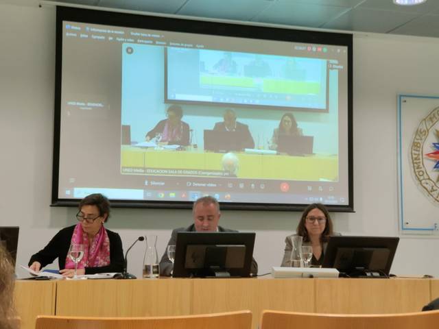 Three people sitting at a desk with laptops in a conference room, under a large projected screen showing the same meeting.