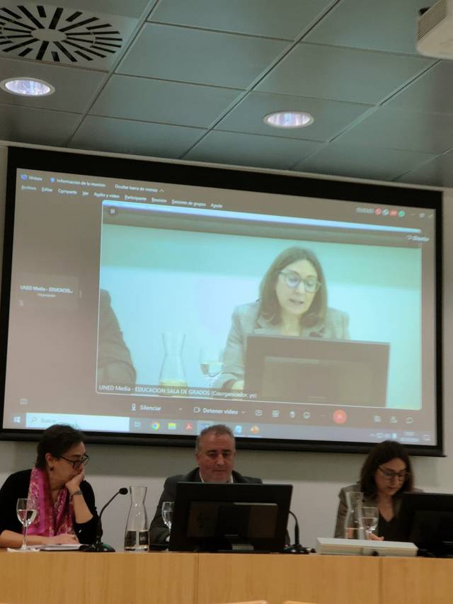 A conference room with people seated at a desk, watching a woman speaking on a large screen.