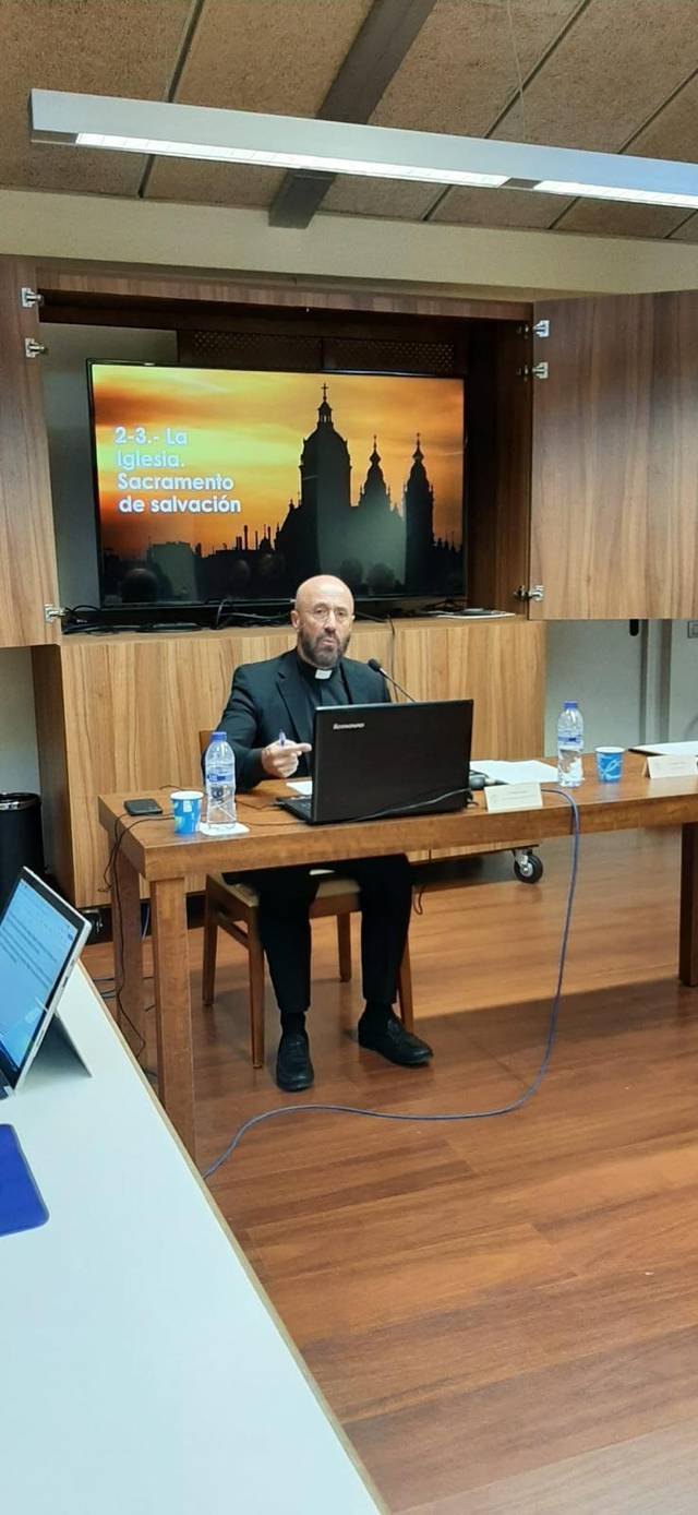 Un hombre está sentado frente a una computadora portátil en una sala de conferencias con un fondo digital que muestra una puesta de sol detrás de siluetas de edificios.