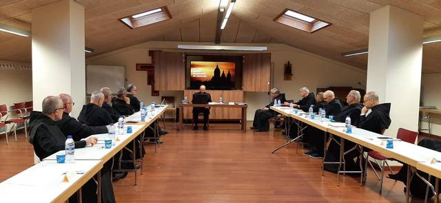 Un grupo de monjes sentados alrededor de largas mesas en una sala de reuniones con una pantalla mostrando una imagen de un atardecer.