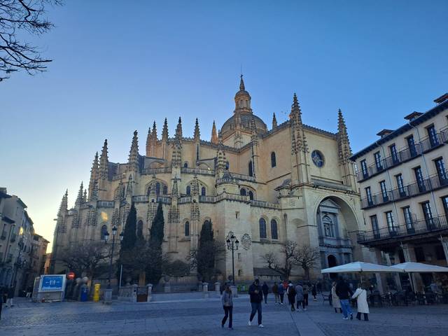 Vista de una catedral gótica en una plaza con algunas personas caminando y edificios alrededor durante el día.