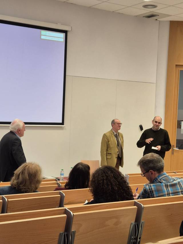 Tres hombres de pie al frente de una sala de conferencias, interactuando con una audiencia en un ambiente académico.