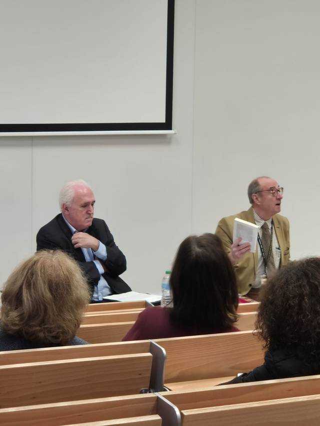 Dos hombres mayores hablando frente a un grupo de personas en un aula universitaria.