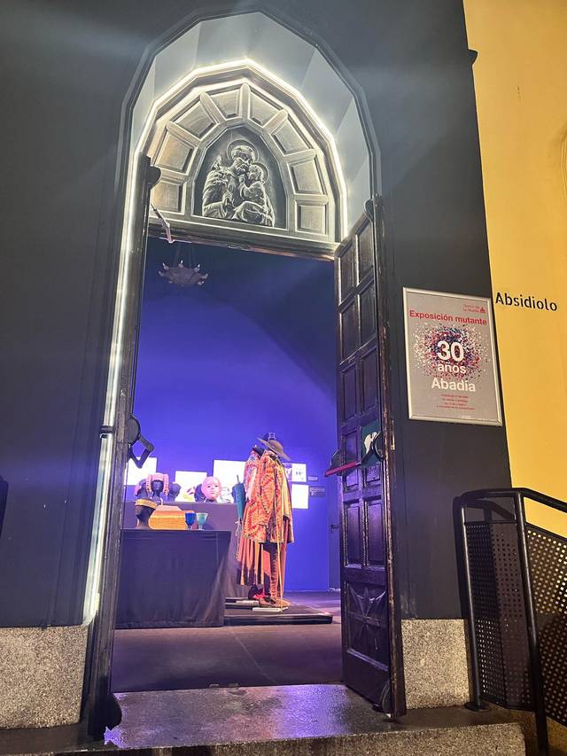 La imagen muestra una entrada a una exposición con un fondo oscuro y una figura vestida con ropa tradicional.