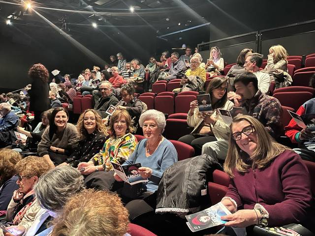 Un grupo de personas se encuentra en un teatro, sentadas y sosteniendo programas del espectáculo.