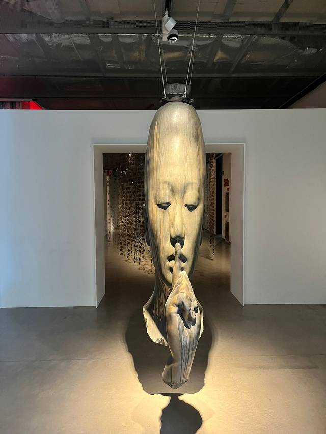 La imagen muestra una escultura suspendida que representa una cara con un dedo en los labios, simbolizando el silencio.