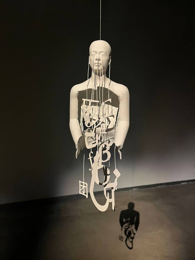 La imagen muestra una escultura suspendida que representa una figura humana hecha de materiales blancos, con un diseño intrincado y un juego de sombras en el fondo.