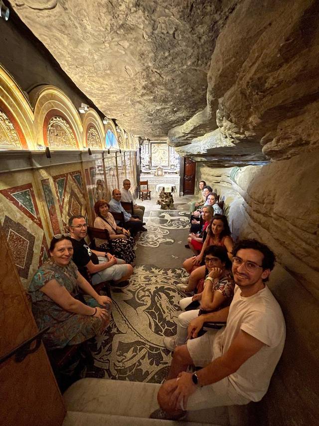 Grupo de personas sentadas en un pasillo decorado con mosaicos en un espacio rocoso.
