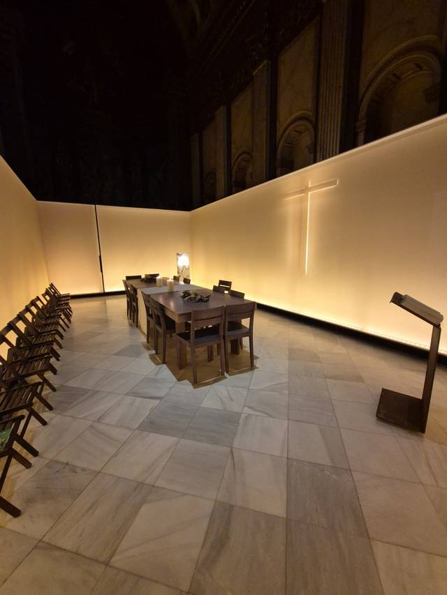 La imagen muestra un interior minimalista con una mesa larga y sillas, iluminado por una suave luz que resalta un gran crucifijo en la pared.