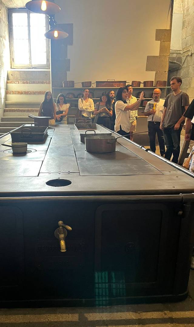 La imagen muestra un gran espacio interior con un objeto metálico, posiblemente una mesa de cocina antigua, y varias personas observando o interactuando entre sí.